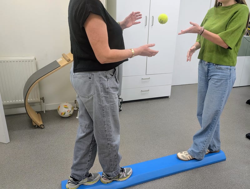 Patiënt oefent evenwicht op een balanceerbalk met een bal, onder begeleiding van de kinesitherapeut